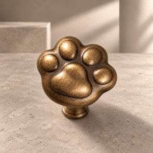 Puede incluir: Un tirador de mueble con forma de huella de animal con un acabado de bronce antiguo desgastado. El herraje presenta una almohadilla central y cuatro almohadillas más pequeñas, todas con una superficie lisa y redondeada.