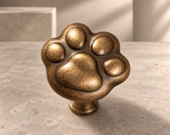 Könnte beinhalten: Ein Möbelknopf in Form eines Tierpfotenabdrucks mit einem antiken, abgenutzten Bronze-Finish. Der Beschlag verfügt über einen zentralen Ballen und vier kleinere Zehenballen, alle mit einer glatten, abgerundeten Oberfläche.