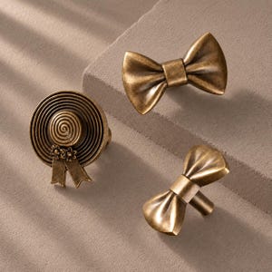 Puede incluir: Tres pomos para armario de metal en tono latón antiguo. Un pomo tiene forma de sombrero de ala ancha con una corona en espiral y dos pequeñas flores en el ala. Los otros dos pomos tienen forma de pajarita con una banda central lisa.