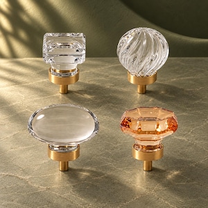 May include: Four cabinet knobs with clear or amber glass tops and brushed brass bases. The set includes a square knob, a spherical knob with a spiral pattern, an oval knob, and an octagonal knob, all featuring a metallic gold-toned finish.
