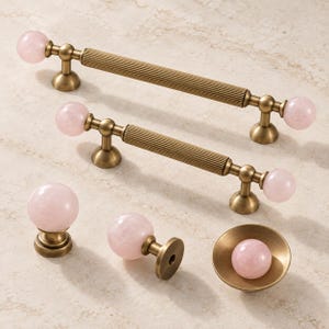 May include: Two brass cabinet pulls with ribbed centers and spherical light pink stone ends, two matching spherical cabinet knobs, and one small brass bowl-shaped knob featuring a central light pink stone sphere.