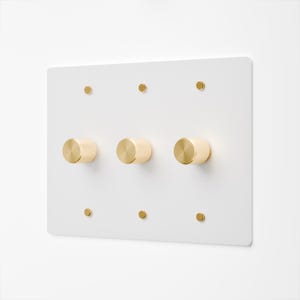 Puede incluir: Una placa de pared rectangular blanca con tres perillas cilíndricas doradas dispuestas horizontalmente en el centro. La placa está asegurada con seis tornillos dorados pequeños, tres posicionados en el borde superior y tres en el inferior.