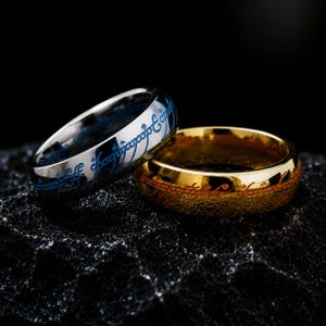 Op de afbeelding: Twee metalen ringen met gegraveerd schrift. De ene ring is zilverkleurig met blauw schrift en de andere is goudkleurig met oranje-rood schrift. Beide ringen hebben een doorlopend patroon van tekens rondom het buitenoppervlak van het metaal.