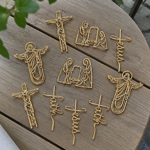Op de afbeelding: Een verzameling goudkleurige metalen paperclips met religieuze ontwerpen, waaronder figuren van Jezus aan het kruis, kerststallen en de in schrijfletters gevormde woorden Jesus en faith, gerangschikt op een lichtbruin houten oppervlak.
