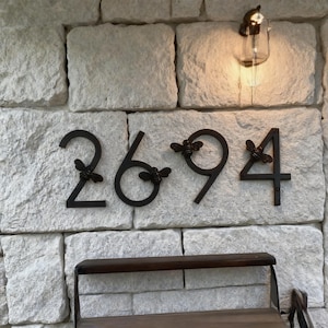 May include: Black metal house numbers 2694 mounted on a light-colored stone wall. Each number is adorned with a small bee accent. A bench sits below the numbers, and a light fixture is above.