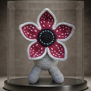 Könnte beinhalten: Gehäkelte Demogorgon-Figur mit einem burgunderfarbenen, blütenartigen Kopf mit weißen Punkten und grauem Rand. Die Mitte ist schwarz mit einer weißen Spirale. Der Körper und die Beine sind grau. Präsentiert in einer transparenten, quadratischen Vitrine.