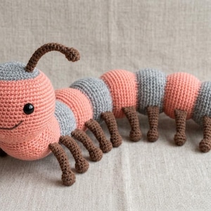 May include: A handmade crochet caterpillar toy in shades of coral, gray, and brown. The caterpillar has a smiling face, segmented body, and multiple brown legs. The toy is designed to be soft and cuddly.