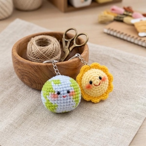 May include: Two handmade crochet keychains: one shaped like the Earth with a smiling face, and the other a yellow sun with a smiling face. The keychains are on silver chains. A wooden bowl with twine and scissors is in the background.