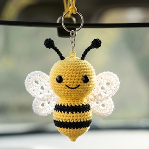 Pode incluir: Um chaveiro de abelha de crochê, amarelo com listras pretas e asas brancas. A abelha tem olhos pretos, antenas e um rosto sorridente. O chaveiro está pendurado em um anel prateado.