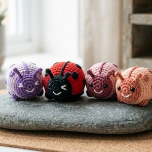 May include: Four handmade crochet insect toys in purple, red, pink, and peach. The toys are shaped like ladybirds and other bugs, with smiling faces and small antennae. They are displayed on a grey stone.