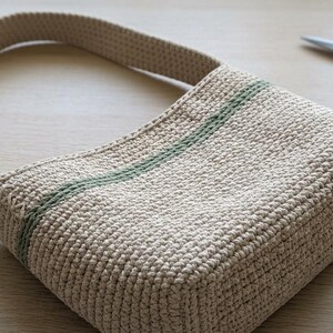 May include: A beige crocheted handbag with a single green stripe detail. The bag has a rounded handle and is sitting on a light wooden surface. A pair of scissors and a crochet hook are also visible.