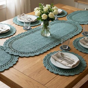 May include: A wooden dining table set for a meal, featuring a teal crocheted table runner, placemats, and coasters. White plates with silverware and folded napkins are set for each place setting. A vase of white roses sits in the center.