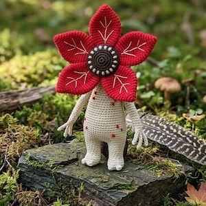 May include: A handmade crochet Demogorgon doll. The doll features a large red flower-like head with white accents, a beige body with red spots, and small arms and legs. The doll is standing on a moss-covered rock.