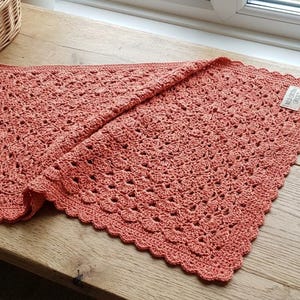 Puede incluir: Manta de ganchillo de color coral con borde festoneado, expuesta sobre una superficie de madera. También se ven una madeja de hilo, un ganchillo y una cesta de hilo. La manta mide aproximadamente 76 cm cuadrados.