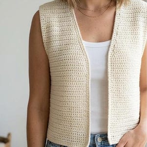 Pode incluir: Colete de crochê sem mangas cor creme com decote em V. O colete aberto é feito de um material tecido texturizado. A modelo está usando uma regata branca e jeans azuis.