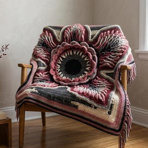 May include: A handmade crochet blanket draped over a wooden chair. The blanket features a central floral design in shades of gray, pink, and cream, with a fringed edge. The chair is positioned near a window, with a vase of dried flowers visible.
