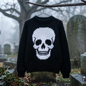 May include: Black knit sweater with a large white skull design on the front. The skull features detailed eye sockets, nose, and teeth. The sweater is hanging on a hanger against a blurred background of a cemetery.