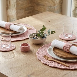 Könnte beinhalten: Tischarrangement mit rosa gehäkelten Tischsets, Untersetzern und Serviettenringen. Jedes Gedeck umfasst eine weiße Serviette, einen Holzteller und Holzbesteck. Eine kleine Schale mit Steinen und Grün ist ebenfalls vorhanden.