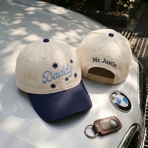 May include: Two cream-colored baseball caps with navy blue brims and navy blue top buttons. One cap features the embroidered word Daddy in light blue script with dark blue star accents. The other cap has the embroidered text Mr. Justin in navy blue.