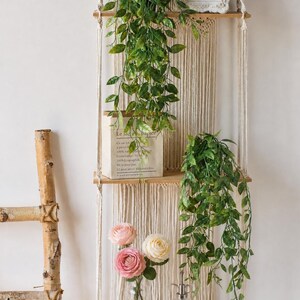 May include: A macrame hanging shelf with two wooden shelves, adorned with faux greenery and a paper bag. The bottom shelf holds a glass vase with pink and cream roses. A small framed picture sits on the top shelf.