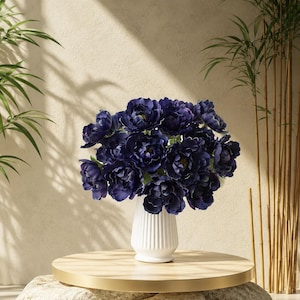 May include: A bouquet of dark blue flowers in a white, ribbed vase. The vase sits on a round, light wood table with a gold rim, supported by large, light-colored stones. Bamboo stalks and leaves are in the background.