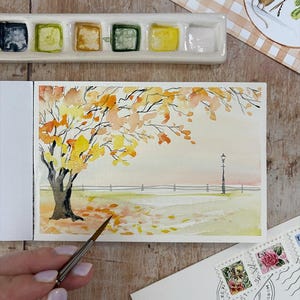 May include: A watercolour painting in a sketchbook depicts an autumn park scene with a large tree featuring yellow and orange leaves, a single black lamppost, and fallen foliage on the ground. A paintbrush tip touches the paper surface.