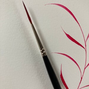 May include: A close-up of a black and silver paintbrush with a red watercolor wash in the background.