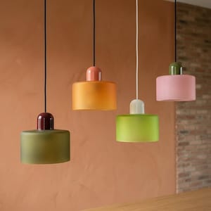 May include: Four pendant lights with cylindrical shades in various colors: olive green, orange, lime green, and pink. Each light has a contrasting top in burgundy, coral, white, and olive green. The lights hang from black and white cords.