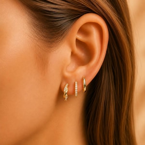 May include: Three gold hoop earrings worn in a single earlobe. The first earring is a twisted hoop with small clear stones, the second is a thin hoop set with a row of clear stones, and the third is a plain, smooth, polished gold hoop earring.