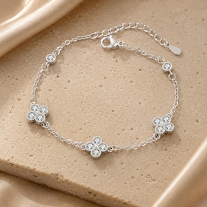 May include: A silver bracelet with a delicate chain design. The bracelet features four clover-shaped charms and several round, clear gemstones. The bracelet is displayed on a neutral-toned surface.
