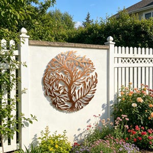 Puede incluir: Un arte mural de metal color cobre con un árbol de la vida está montado en una pared blanca. El diseño del árbol es circular, con intrincadas ramas y hojas. La pared forma parte de un jardín con una valla blanca y flores coloridas.