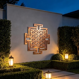 May include: A large, square, copper-coloured metal wall art piece featuring a geometric flower of life pattern within a structured, angular frame. The piece is mounted on a smooth, light-coloured exterior wall and is illuminated by warm outdoor lighting.