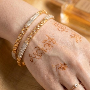 Puede incluir: Dos brazaletes dorados con detalles de diamantes se muestran en una mano con diseños de henna. También se ve un anillo de oro con un solo diamante. Las joyas se presentan sobre un fondo cálido.