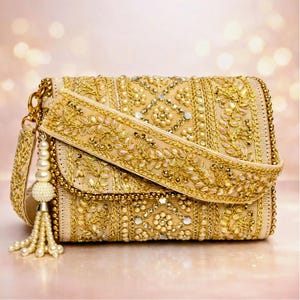 Puede incluir: Un bolso de mano rectangular dorado con intrincados bordados y adornos. El bolso presenta una correa larga y una borla decorativa con cuentas perladas. El diseño incluye patrones florales y pequeños acentos brillantes, creando un aspecto lujoso.