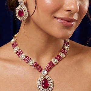 Puede incluir: Un conjunto de collar y pendientes de color dorado con piedras rojas rubí en forma de lágrima y piedras claras de acento. El collar tiene un colgante central con una gran piedra roja rubí. Los pendientes reflejan el diseño del colgante.