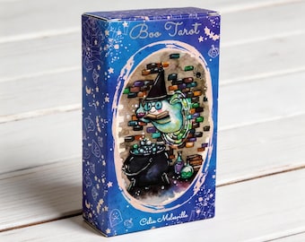 May include: A rectangular deck box for Boo Tarot cards by Cilia Melesville. The blue box features a central illustration of a ghost wearing a witch hat and glasses, reading a book above a black cauldron, set against a brick wall background.