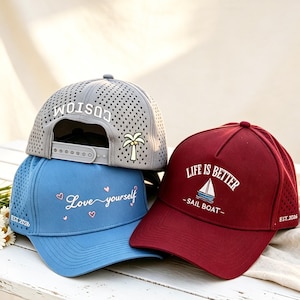 Könnte beinhalten: Drei Baseballkappen in Grau, Blau und Kastanienbraun. Die graue Kappe zeigt eine Palme und das Wort CUSTOM. Die blaue Kappe trägt den Schriftzug Love yourself mit Herzen. Die kastanienbraune Kappe zeigt LIFE IS BETTER SAIL BOAT und EST. 2026.