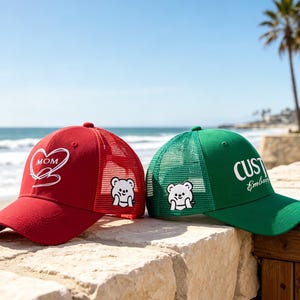 Könnte beinhalten: Eine rote Trucker-Cap mit einem weißen Herz-Motiv und dem Text MOM sowie eine grüne Trucker-Cap mit dem Text CUSTOM Embroidery. Beide Caps haben ein weißes Bären-Motiv an der Seite, Netz-Einsätze am Hinterkopf und einen gebogenen Schirm.