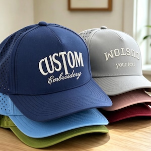 May include: A stack of five baseball caps in navy, light blue, olive green, light gray, and maroon. The navy cap features white embroidered text reading CUSTOM Embroidery, while the light gray cap features white text reading CUSTOM your text.