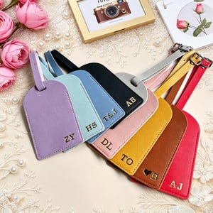 May include: A selection of personalised luggage tags in shades such as lavender, light blue, black, pink, yellow, brown, and red. Each tag displays gold lettering with initials or a heart and initial. The tags are arranged on a decorative surface.