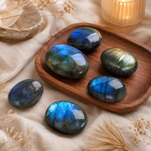 May include: Six polished, oval-shaped labradorite stones with smooth surfaces. The stones exhibit dark gray and charcoal tones with iridescent flashes of vibrant blue and golden yellow light reflecting across their polished, rounded surfaces.