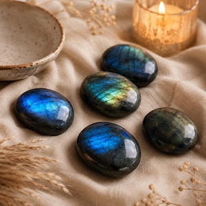 May include: Five polished, oval-shaped labradorite stones featuring dark grey surfaces with iridescent flashes of blue and gold light. The stones are arranged on a textured, light-colored fabric surface near a ceramic bowl and a lit candle.