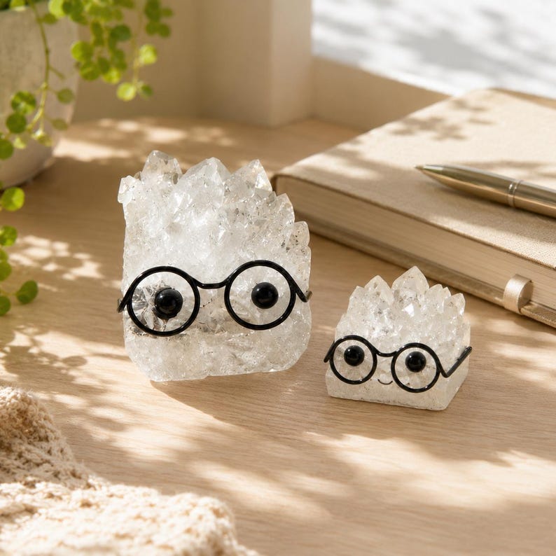 May include: Two clear, jagged quartz crystal clusters shaped like figures, each wearing small black wire-rimmed glasses with round frames. The larger crystal is on the left, and the smaller crystal is on the right, both featuring smooth, flat bases.