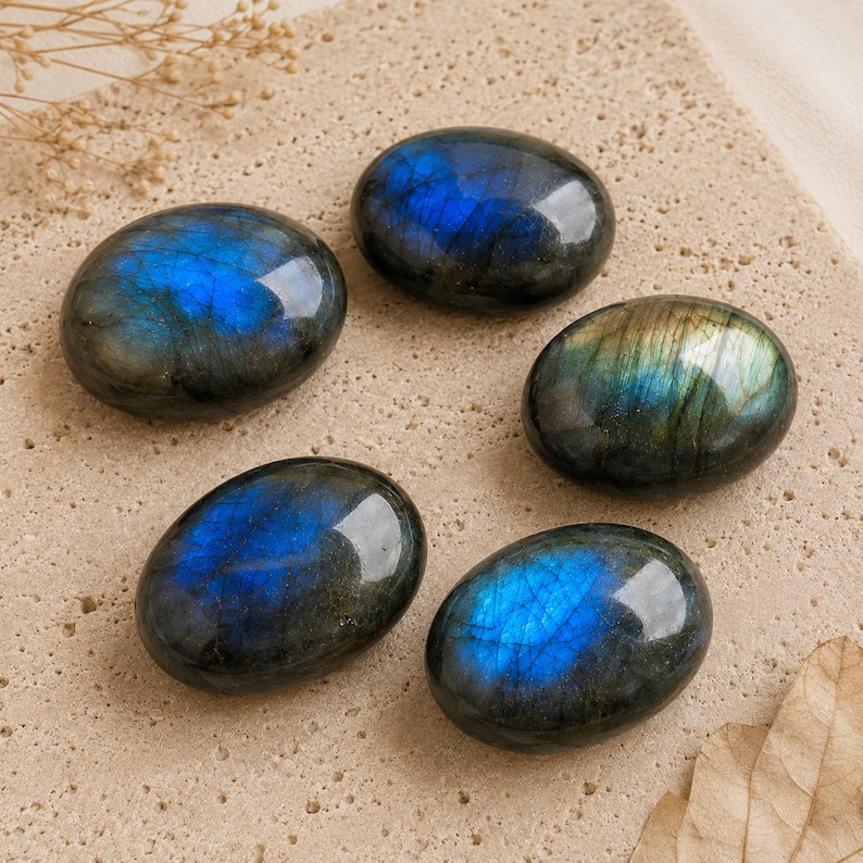 May include: Five polished oval labradorite stones featuring dark gray bases with iridescent blue and gold flashes. Each stone has a smooth, rounded surface that reflects light, highlighting the natural mineral inclusions and color variations.
