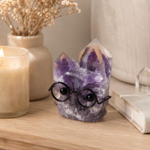 May include: A cluster of deep purple amethyst crystals shaped into a figure wearing black wire-rimmed glasses. The crystal formation features multiple pointed peaks and a rough, faceted texture, with the black glasses resting across the front.
