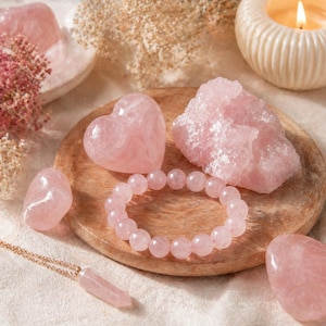 May include: A collection of rose quartz crystals and jewelry. The arrangement includes a heart-shaped stone, a bracelet, a pendant necklace, and raw crystal formations. A lit candle and dried flowers are in the background. The stones are various shades of pink.