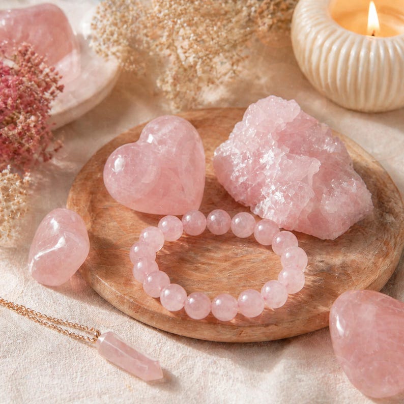 May include: A collection of rose quartz crystals and jewelry. The arrangement includes a heart-shaped stone, a bracelet, a pendant necklace, and raw crystal formations. A lit candle and dried flowers are in the background. The stones are various shades of pink.