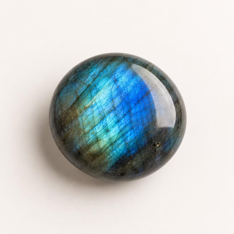 May include: A polished, circular labradorite gemstone featuring a smooth, rounded surface. The stone displays a vibrant, iridescent play of color, shifting between deep electric blue, teal, and dark charcoal gray tones with visible internal fractures.