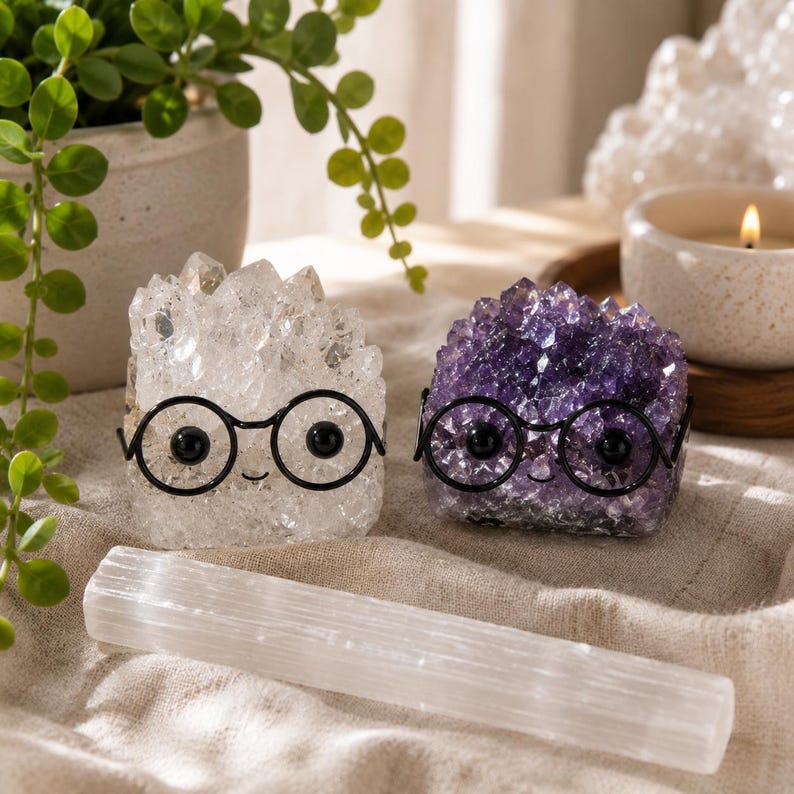 May include: Two crystal clusters, one clear and one purple, each featuring black wire-rimmed glasses and small black eyes. A long, rectangular white selenite wand lies horizontally in front of the two crystal figures on a textured fabric.