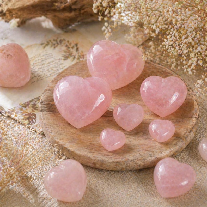 May include: A collection of polished rose quartz hearts in various sizes. The hearts range in color from pale to medium pink and are arranged on a wooden plate and a sandy surface. The image has a soft, natural aesthetic.